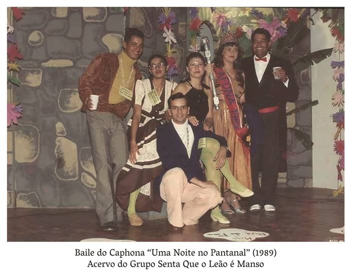 4 g.  Foto colorida Baile da Caphona Uma noite no Pantanal (1989) Acervo do Grupo senta que o Leão é manso. Três casais de homens e mulheres estão trajando roupas coloridas. Esses casais estão encostados em uma parede cinza e branca. Uma mulher está usando um vestido dourado e uma coroa, usa também uma faixa onde aparece escrito: Cafona 1989i. Realização: Academia Planaltinense de Letras, Artes e Ciências (APLAC), pelo Ecomuseu Pedra Fundamental e pelo Coletivo Nativo Audiodescrição produzida  pelo Instituto de Promoção das Pessoas com Deficiência Visual Audiodescritora: Elma Lúcia Rodrigues Consultor: Fernando Rodrigues Este projeto é promovido com recursos do Fundo de Apoio a Cultura do DF